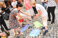 Ein Junge und eine Frau legen auf einem Steinfußboden ein Arrangement für Fotoaufnahmen zurecht. Das Arrangement besteht aus einer Tafel in der Mitte auf der "Kalendergänger" steht und drei Sprechblasen ringsherum, auf denen Statements zum Thema Demokratie stehen.