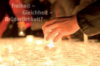 Ein Meer von Kerzen mit Händen, die neue Kerzen hinzustellen. Mit Schriftzug "Freiheit - Gleichheit - Brüderlichkeit"