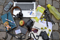 Auf dem Boden liegen ein Laptop, ein Handy, ein Notizbuch, ein Fotoapparat, ein Objektiv, Kopfhörer, eine Sonnenbrille, ein Stadtplan, eine Wasserflasche, ein Stativ, ein Stift, ein kleiner Plüschlöwe, Bonbons, eine Fahrkarte, ein Bibliotheksausweis