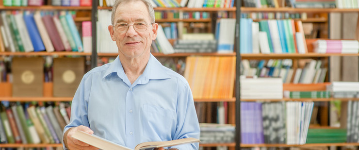 Ein älterer Mann steht mit einem aufgeschlagenes Buch in den Händen vor einem Bücherregal