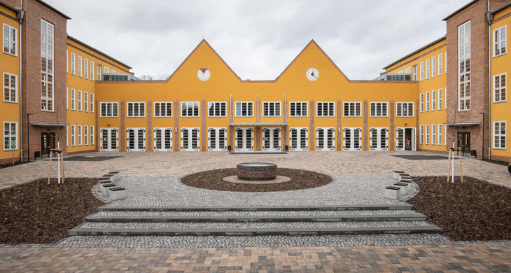Auf dem Bild erkennt man ein Schulgebäude mit Orange-gelber Fassade. Im Vordergrund ist der Schulhof zu sehen.