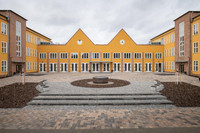 Auf dem Bild erkennt man ein Schulgebäude mit Orange-gelber Fassade. Im Vordergrund ist der Schulhof zu sehen. 