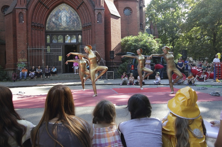 Tänzerinnen in goldenen Ganzkörperkostümen performen vor einer Kirche. Um sie herum sitzt und steht Publikum.