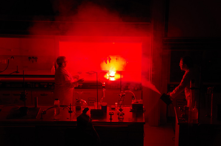 Gelbe Flamme in einem Chemielabor, die den Raum in rotes Licht taucht. Daneben zwei Menschen im Laborkittel