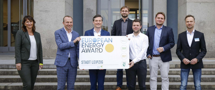 Eine Frau und sechs Männer stehen auf einer Treppe und halten ein Schild in die Kamera, auf dem "European Energy Award" steht.
