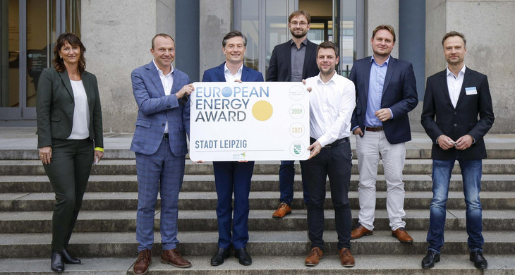 Eine Frau und sechs Männer stehen auf einer Treppe und halten ein Schild in die Kamera, auf dem "European Energy Award" steht.