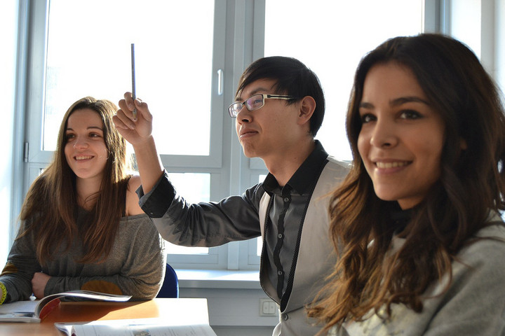 Drei Studenten in einem Zimmer. Ein Student meldet sich mit einem Stift in der Hand.