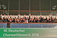 Mitglieder des Kammerchor Josquin stehen hinter einem Geländer und winken. Unter ihnen ist ein Banner: 10. Deutscher Chorwettbewerb 2018.