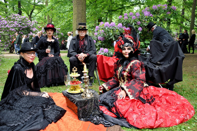 Eine Frau in schwarzem Kleid und ein Mann in schwarzem Anzug und mit HUt sitzen auf Stühlen im Park, davor sitzen auf einer Decke zwei weitere Frauen, eine im schwarzen Kleid, die andere in einem roten Kleid und mit aufwendigem Kopfschmuck.