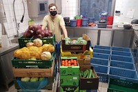 Zoopfleger mit Mund-Nasen-Bedeckung steht in einer Küche hinter Kisten mit verschiedenen Obst und Gemüsesorten