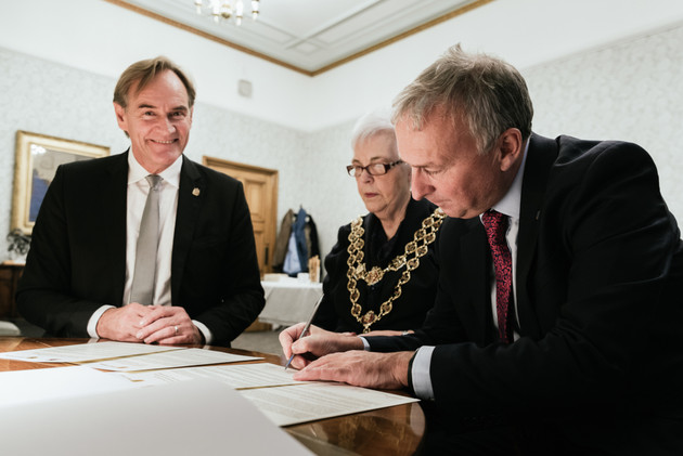 drei Personen stehen an einem Tisch zur Unterzeichnung eines Vertrages