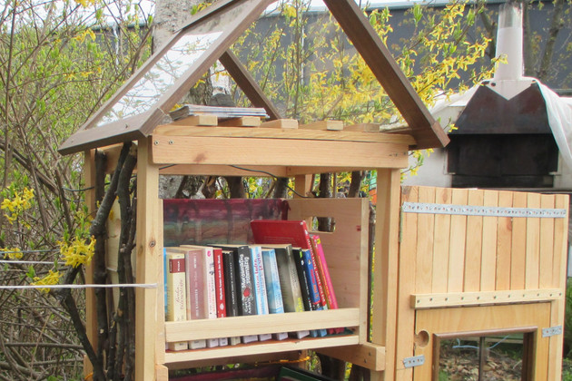 In einem Holzregal mit einem Dach in einem Garten stehen Kinderbücher.