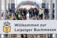 Blick in die gläserne Halle auf der Neuen Messe mit vielen Besuchern. Über der Treppe hängt ein banner mit der Aufschrift "Willkommen zur Leipziger Buchmesse".