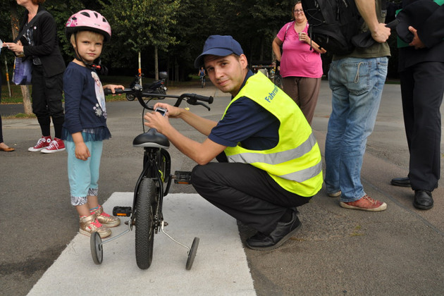 Ein Mann mit gelb leuchtender Weste registriert eine Kinderfahrrad mit Stützrädern. Eine Mädchen mit Fahrradhelm auf dem Kopf steht daneben.