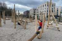 Ein Spielplatz aus vielen Holzstämmen mit Seilen, Tauen und Netzen vor den Wohngebäuden auf dem Lindenauer Hafen. Ein Kind hangelt sich darauf entlang.
