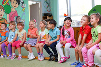 Kindergartenkinder sitzen in einer Reihe auf einer Bank in einem bunten Raum.